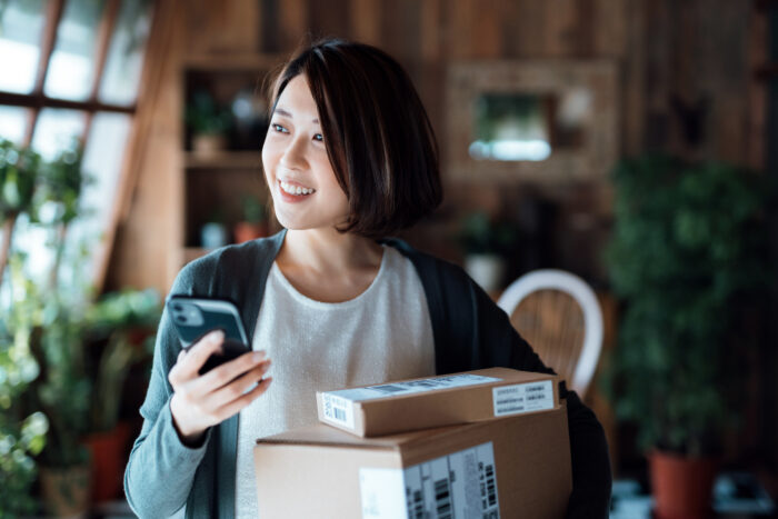 Smiling young Asian woman with a smartphone, receiving parcels with home delivery service.