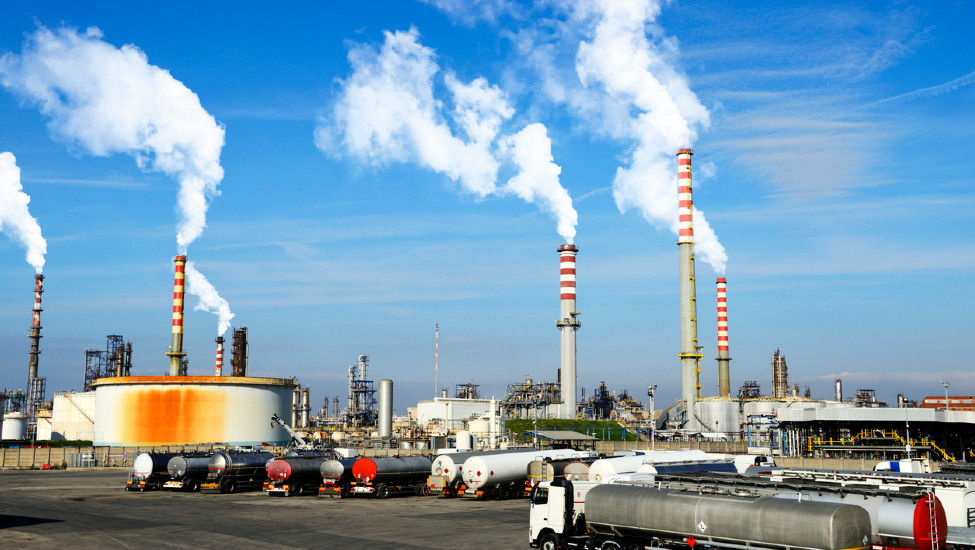 A chemical plant made up of various industrial buildings with six smokestacks emitting white smoke sit in front of several tanker trucks parked in the foreground.