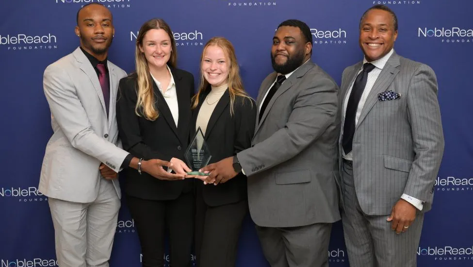 Dressed in business attire, Howard University MBA Exclusive Case Competition team members Antonio Terrell, Emma “Em” Strieter, Lillia “Lily” Hendrickson, Anthony “Tony” Gunn and team advisor Randy V. Bradley pose with their competition trophy.