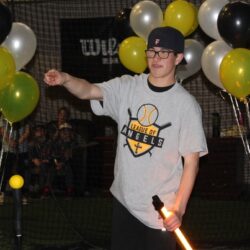Young man wearing glasses, backward baseball cap and League of Angels T-shirt, holding a light-up sword in one hand and pointing with the other. Balloons in background.