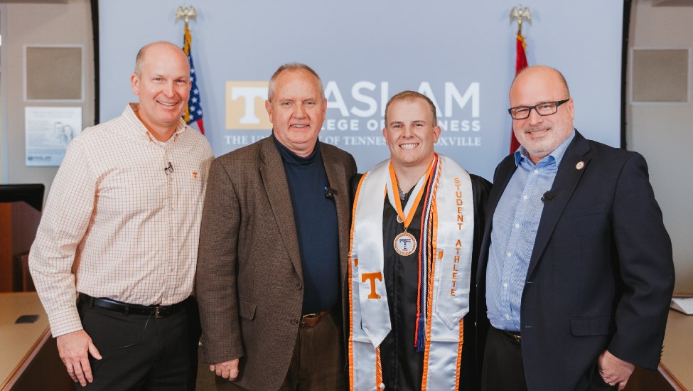Russell Crook, Lane Morris, Christian Weaver in graduation regalia, and George Drinnon pose with American flag and Haslam logo on wall in background