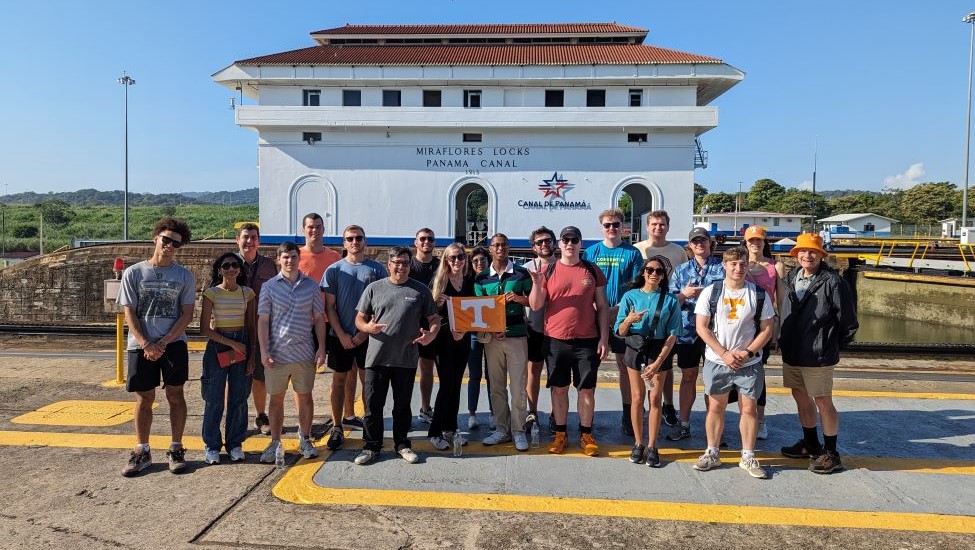In this picture taken on Thursday, January 11, 2024 at the Panama Canal's Miraflores Lock facility, a multiethnic group of students, Professor Rodrigues and a tour guide, all dressed in casual, summertime attire, stand in front of one of the Miraflores Locks buildings, with two students in the center of the group holding a small Power T flag. The business activity, one of the highlights of the study abroad program, encompassed a half day tour of the Panama Canal with a visit to the Pacific Entrance of the canal and a stop at the Miraflores Lock Center, which includes an overview of the Panama Canal’s history, its importance to Panama and its role today.
