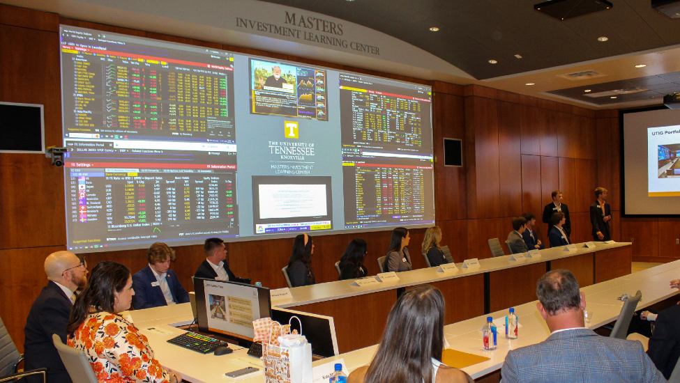 Interior of Masters Investment Learning Center. People seated at conference table with large screen showing financial market info on wall.