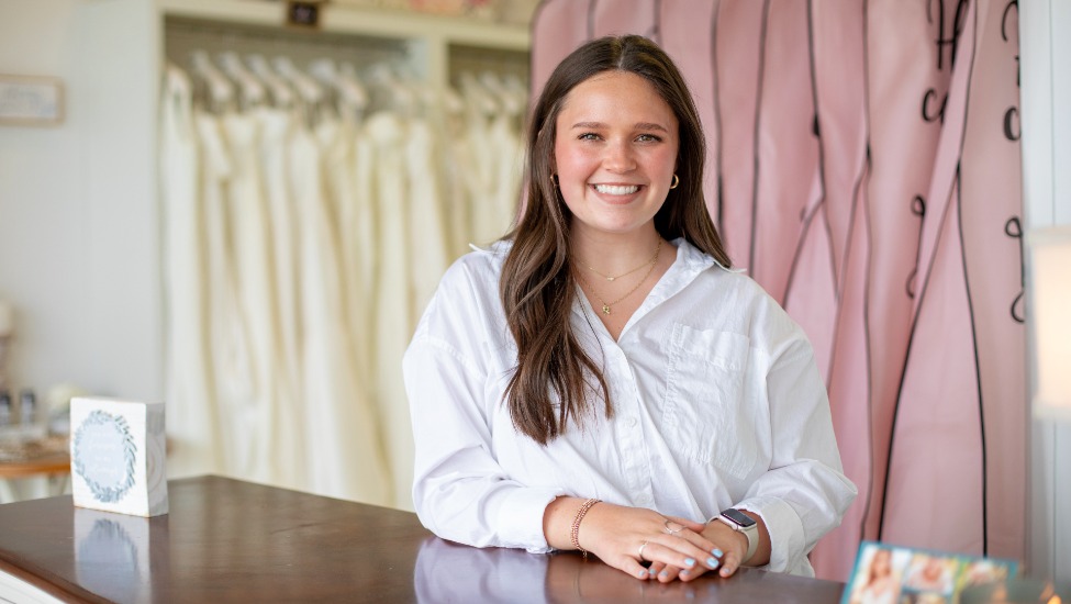 Rheagan Haynes stands behind the counter of her bridal shop, wearing a white long-sleeve shirt and smiling at the camera