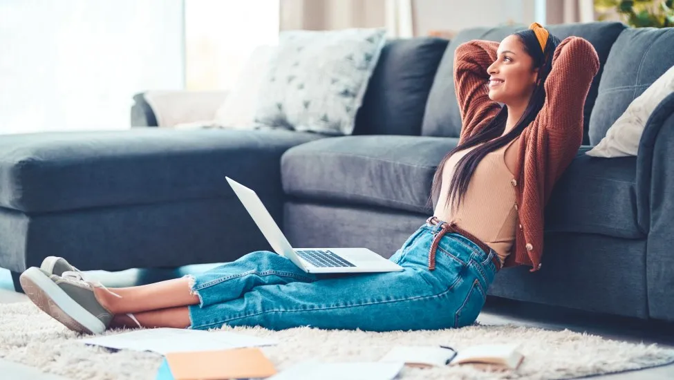 Young woman sitting on floor with laptop on lap, leaning back on couch with relaxed smile on her face.