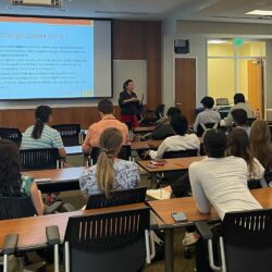 Photo from back of classroom shows students attending to female lecturer speaking.