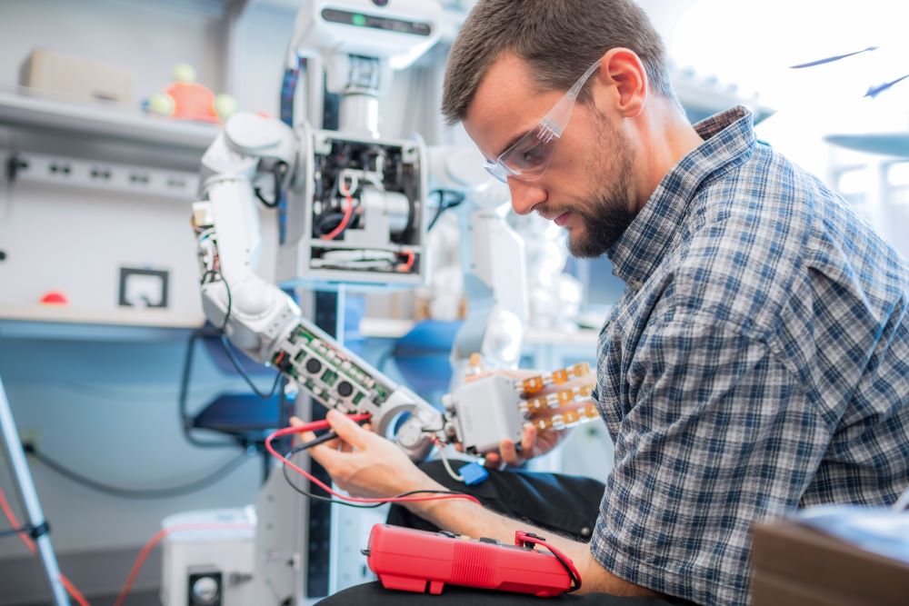 Male worker in plaid shirt and safety goggles performs robot-assisted labor.