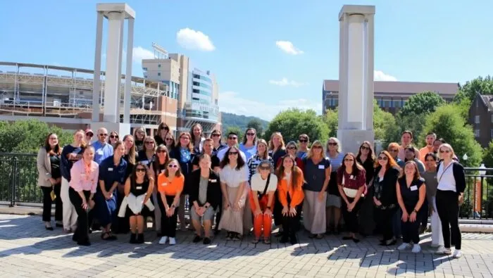 Large group of BisNet 2024 participants dressed in casual clothing - some in UT orange - pose on UT's pedestrian bridge with Neyland Stadium in the background