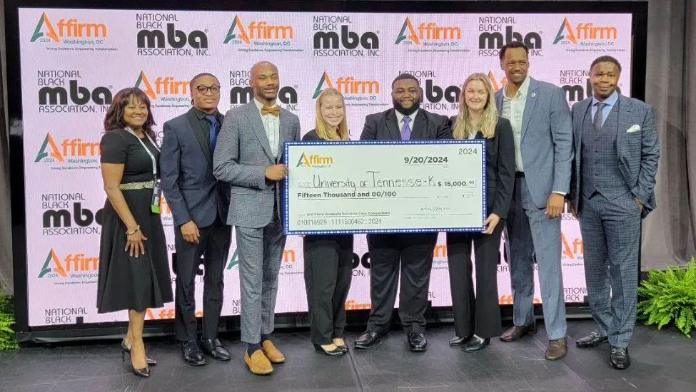 Members of Haslam's MBA case competition team, dressed in business attire, pose with a giant 2nd-place check, with members of the NBMBAA panel