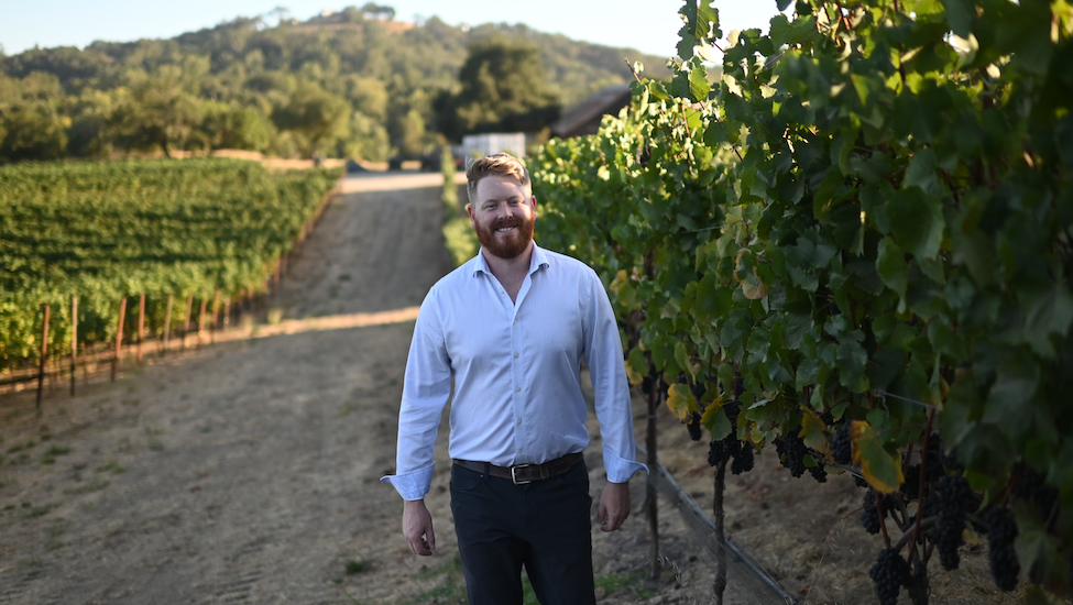 Charles Dick walks beside a row in a California vineyard. He has red hair and a red beard and mustache. He is wearing a light, striped button-up shirt and dark pants.