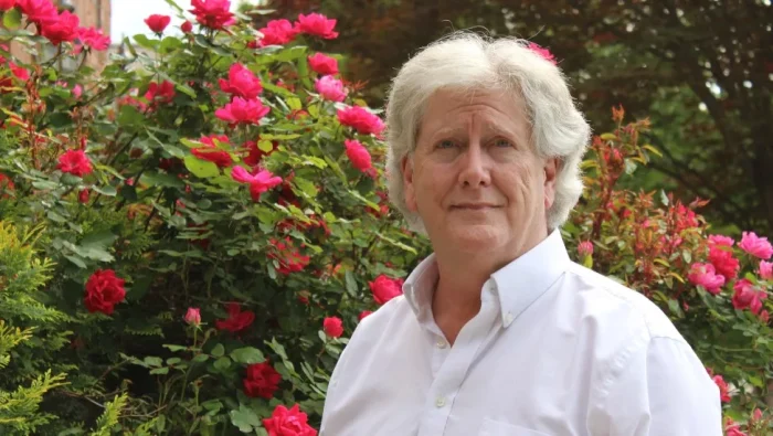 Mark Collins Smiling Wearing a White Collared Shirt in a Background of Red and Pink Roses