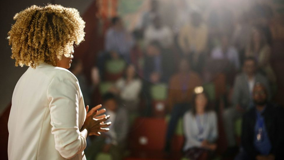 Three-quarter behind-the-back view of a black woman with blondish frizzy hair in a white business coat addressing an audience in a blurred, dim background