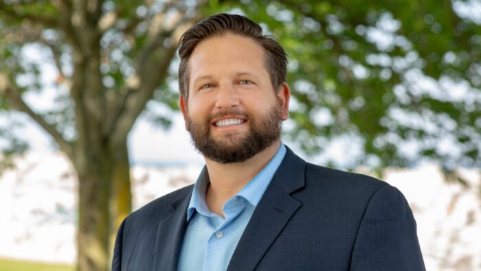 Portrait image of Patrick Booth wearing a sports coat and blue dress shirt standing outside with sunlit trees in the background