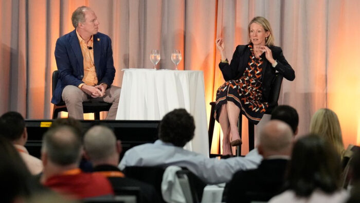 Seated on a stage on either side of a small table draped in a white tablecloth with two glasses of water on it, Tom Goldsby, dressed in light brown slacks, an orange-and-white checkered shirt and blue blazer, listens intently as Marianne Wanamaker in a black dress crisscrossed with an abstract orange-white-pattern and a black blazer, holds forth on tariffs.