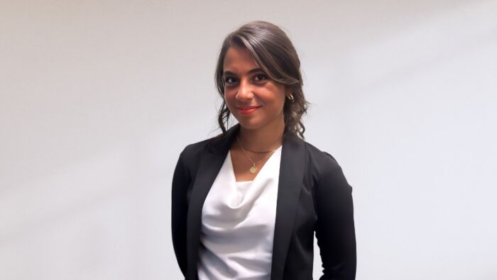 Saba Toski in a white blouse and dark suit jacket stands in front of a gray-white backdrop.