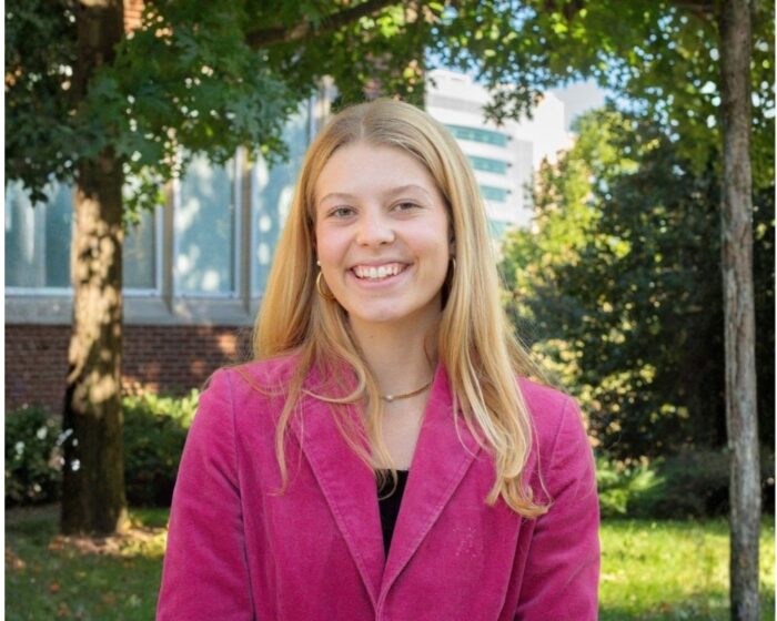 Zoe Shockites, in a mauve jack and black top, stands in an outdoor setting with sunlit trees and shrubs in the background.