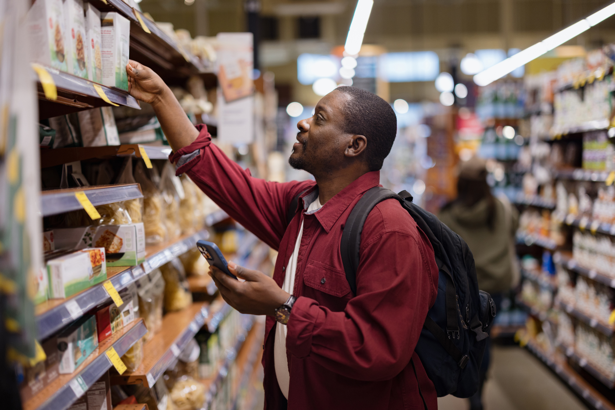 Adult man reaching for a product on a shelf, comparing prices and checking a shopping list on his smartphone in a grocery store